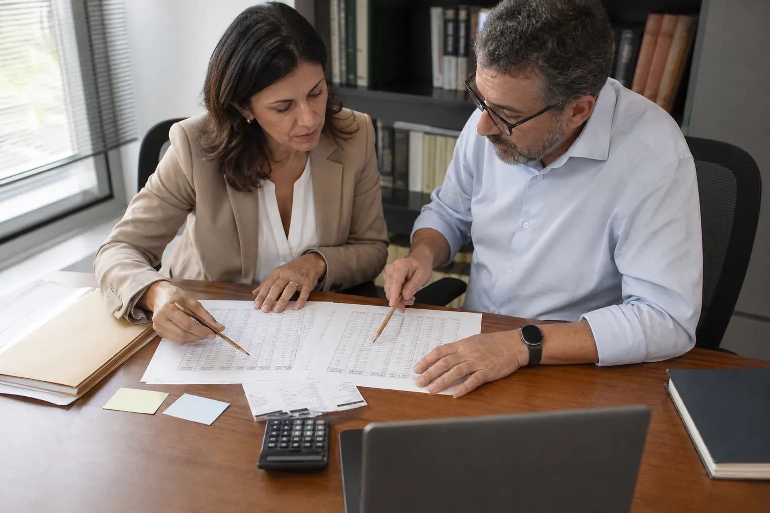 Advogada e analista conferem uma tabela de cálculo e uma calculadora em uma mesa de escritório.